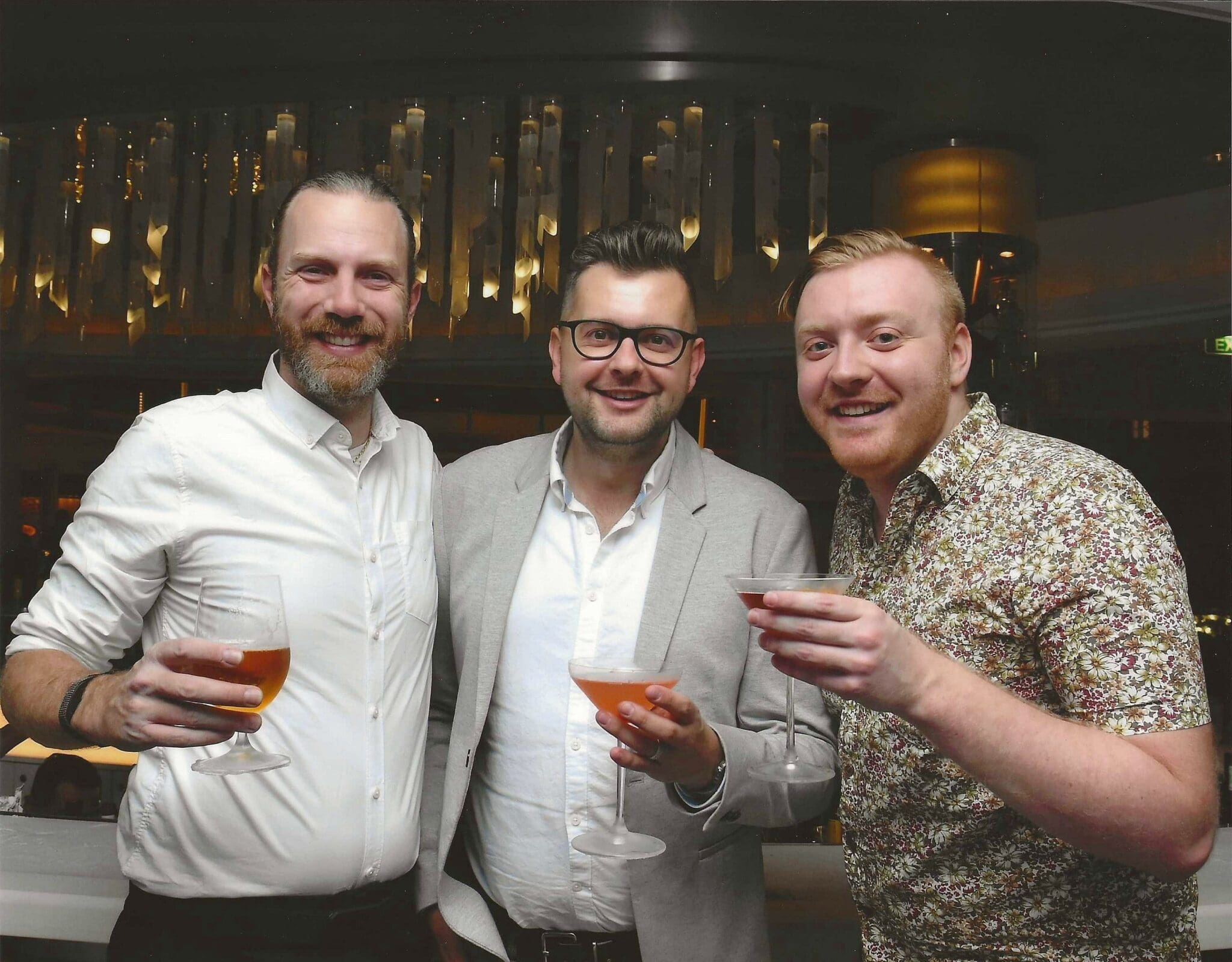 Three men are smiling while holding drinks at a bar.